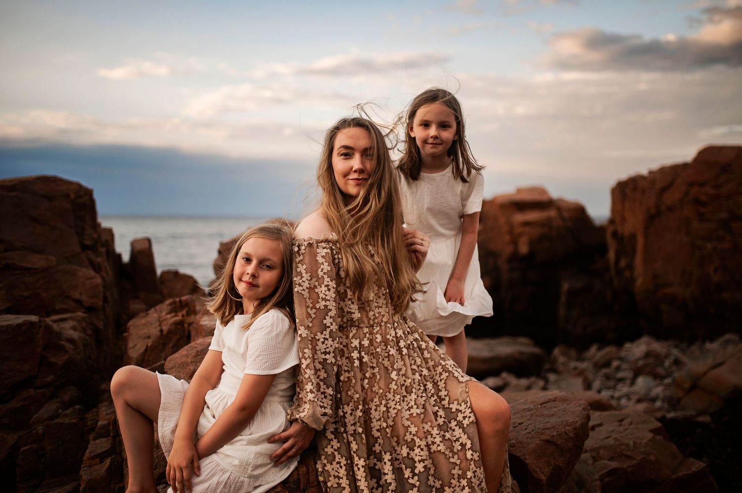 People in flowy dresses pose atop rocky coastal formations at sunset.
