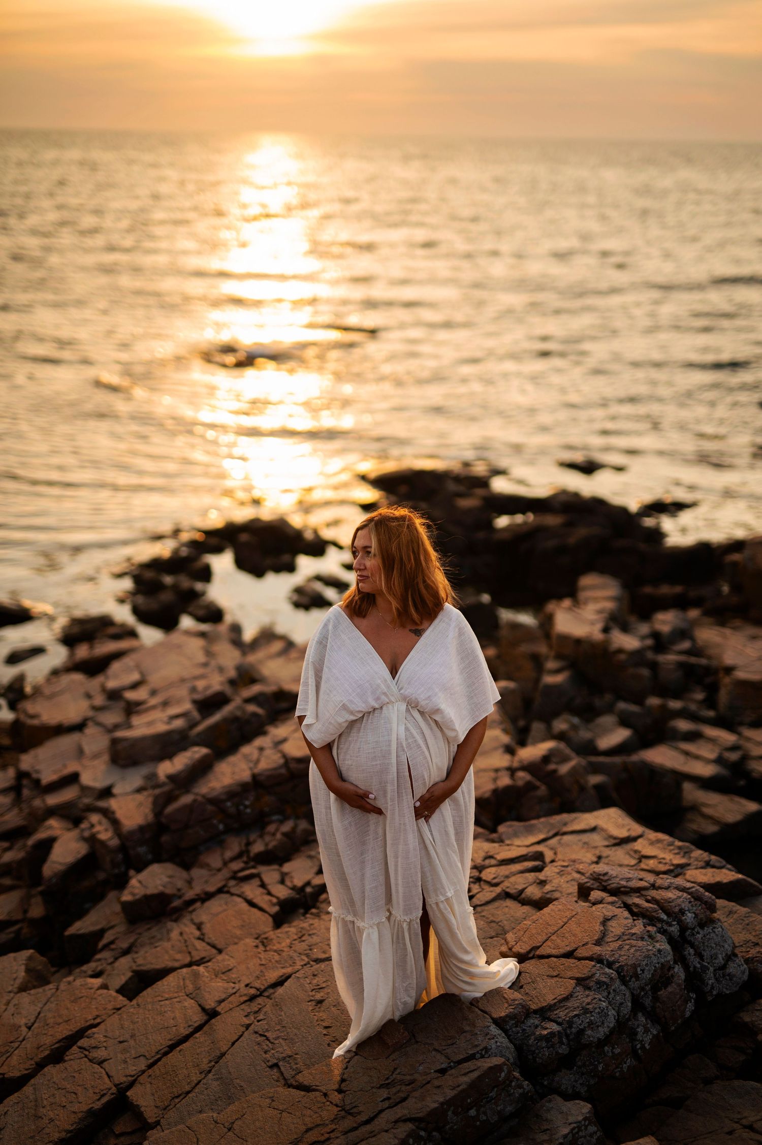 Woman in white dress stands on rocky shore during stunning ocean sunset.