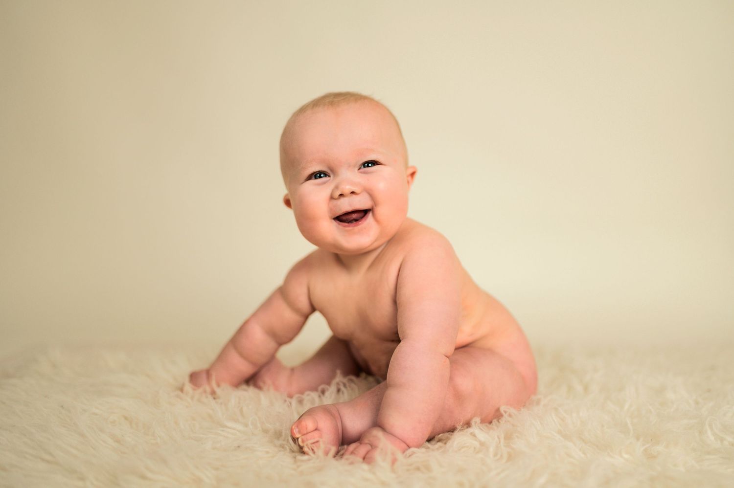 Baby smiling while crawling on soft white fur rug against beige background.