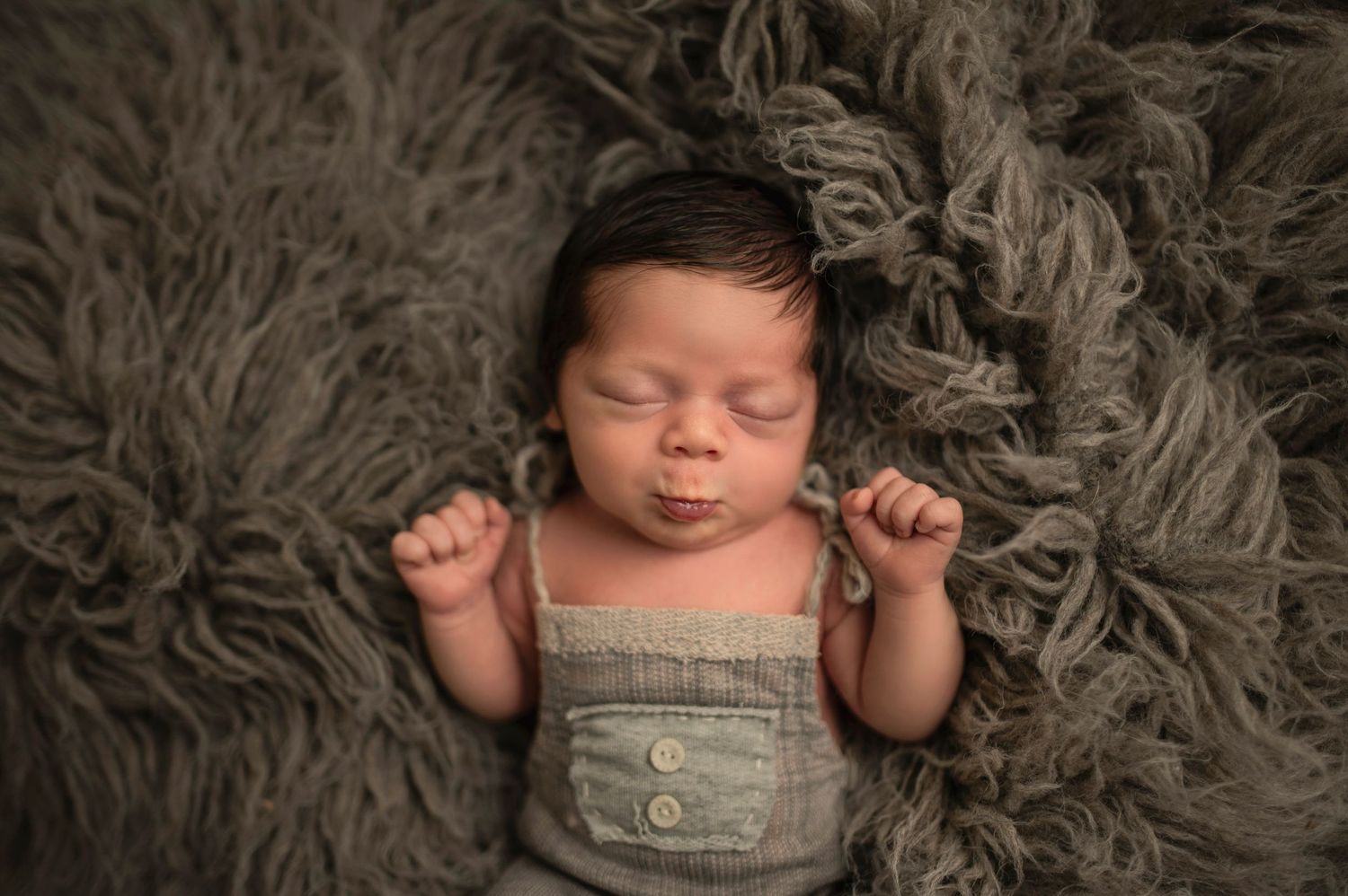 Sleeping newborn wearing overalls on gray fluffy blanket.