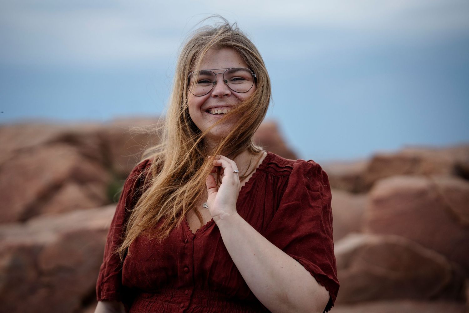 Smiling blonde woman with glasses in red dress posing near red rock formations outdoors.