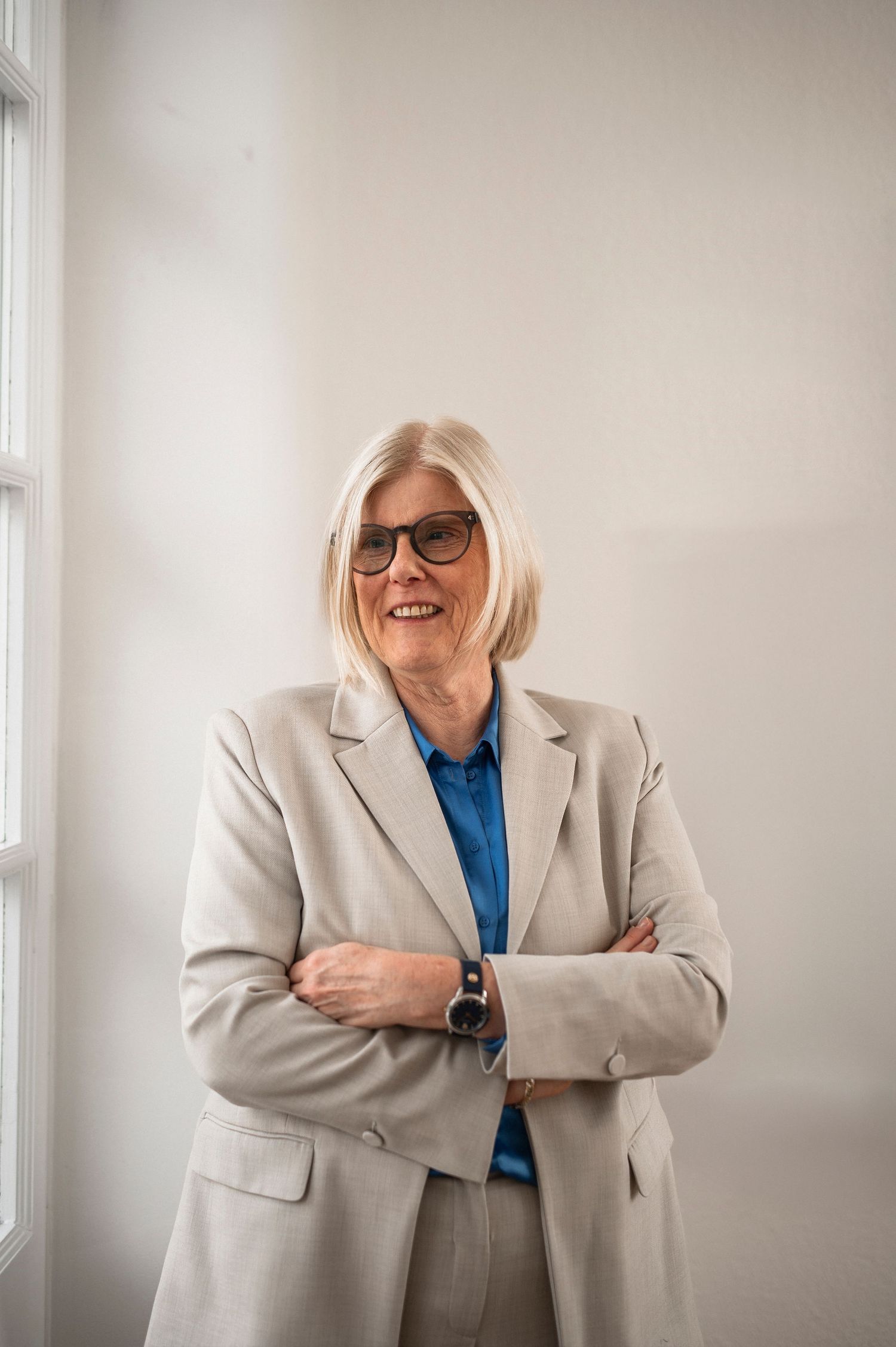 Confident senior businesswoman in gray blazer standing with arms crossed smiling in bright office.