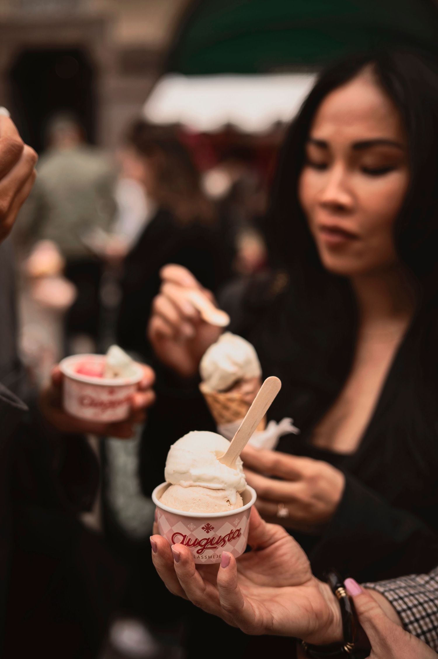 Woman holding an Augusta branded ice cream cup at an outdoor market, tasting gelato with a wooden spoon.