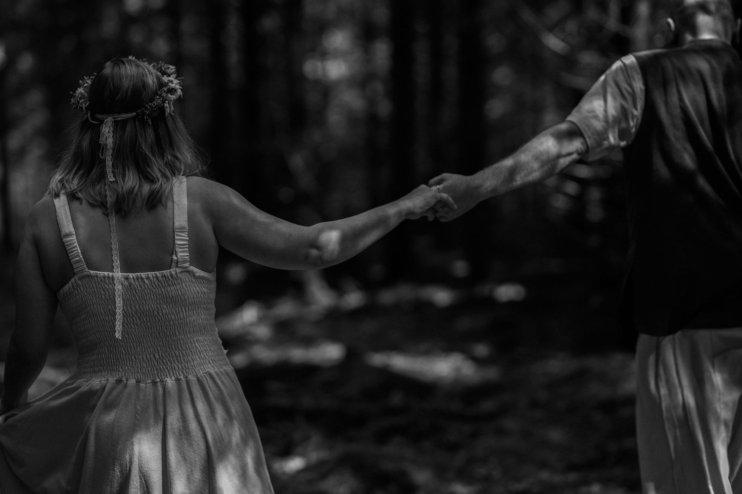 Black and white photo of girl in floral crown holding adult hand in forest.