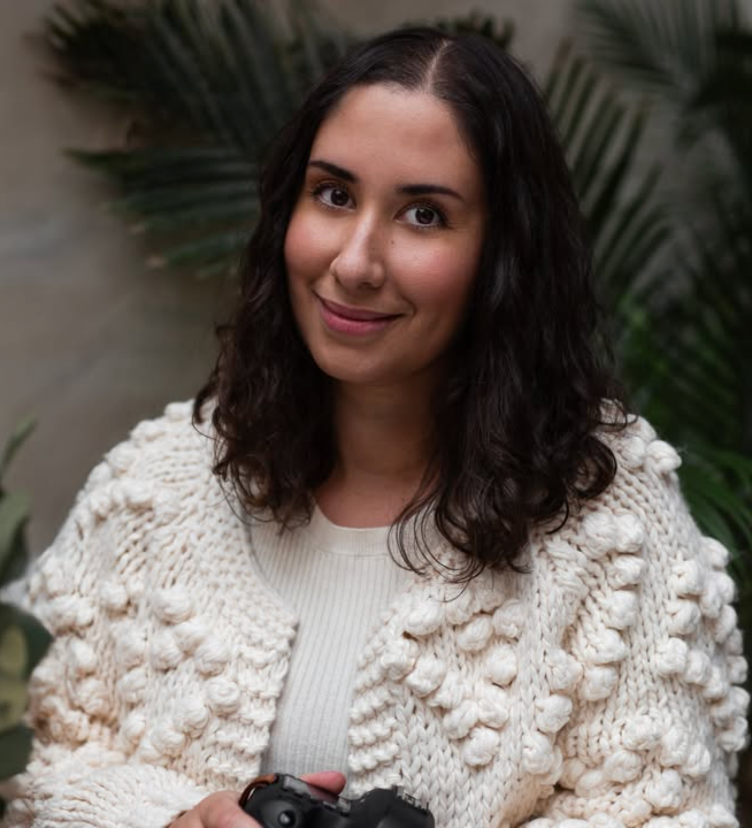 Woman with dark curly hair holding camera wearing white knit cardigan indoors.