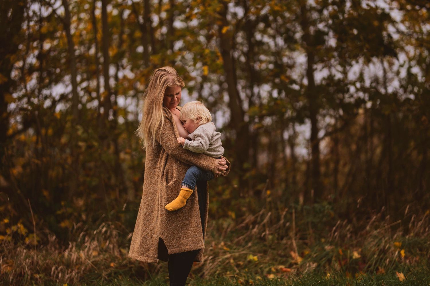 Person in brown coat holding small child with yellow boots in autumn forest setting.