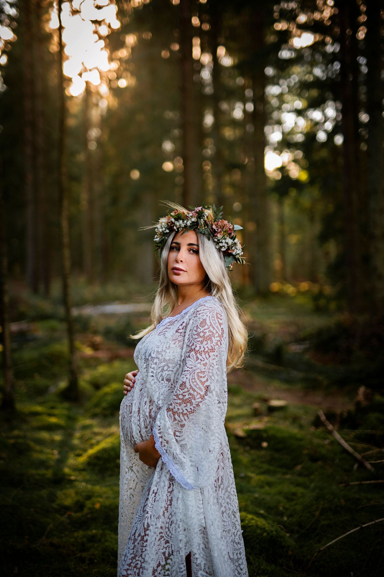 Enchanted forest maternity portrait with flower crown and lace dress amid pine trees.