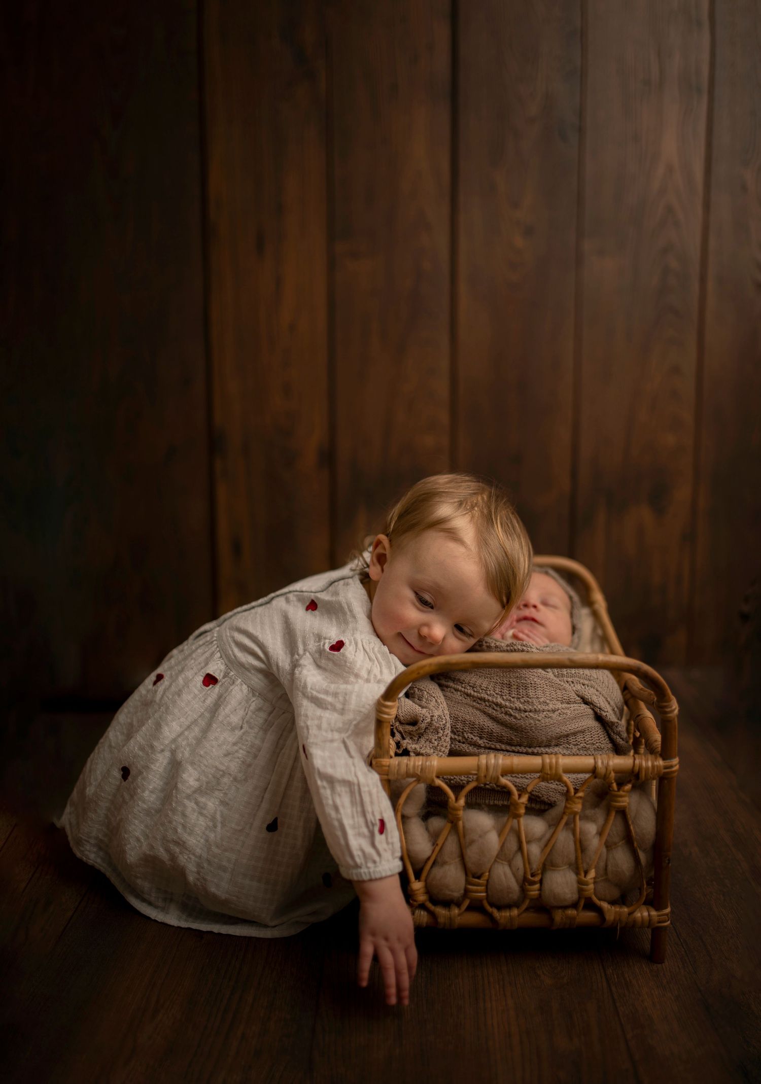 Tender sibling moment with toddler resting head on baby in vintage wicker basket.