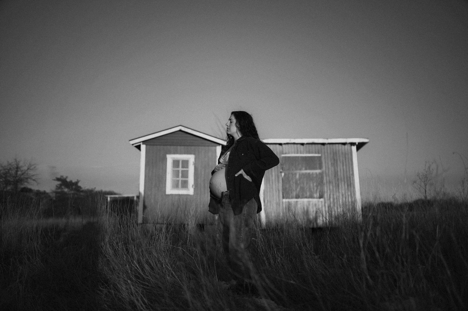 Maternity portrait in black and white with wooden shed and tall grass.