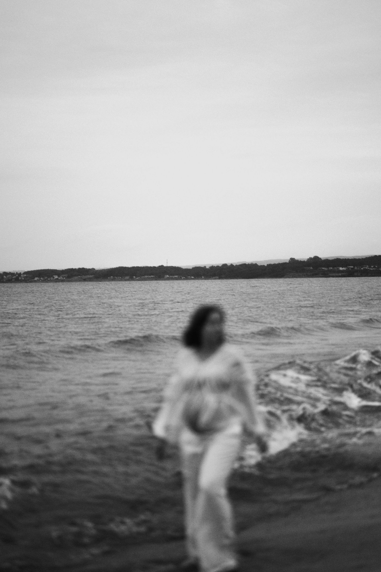 Artistic black and white photo of a figure in white clothing walking along a misty beach shoreline.