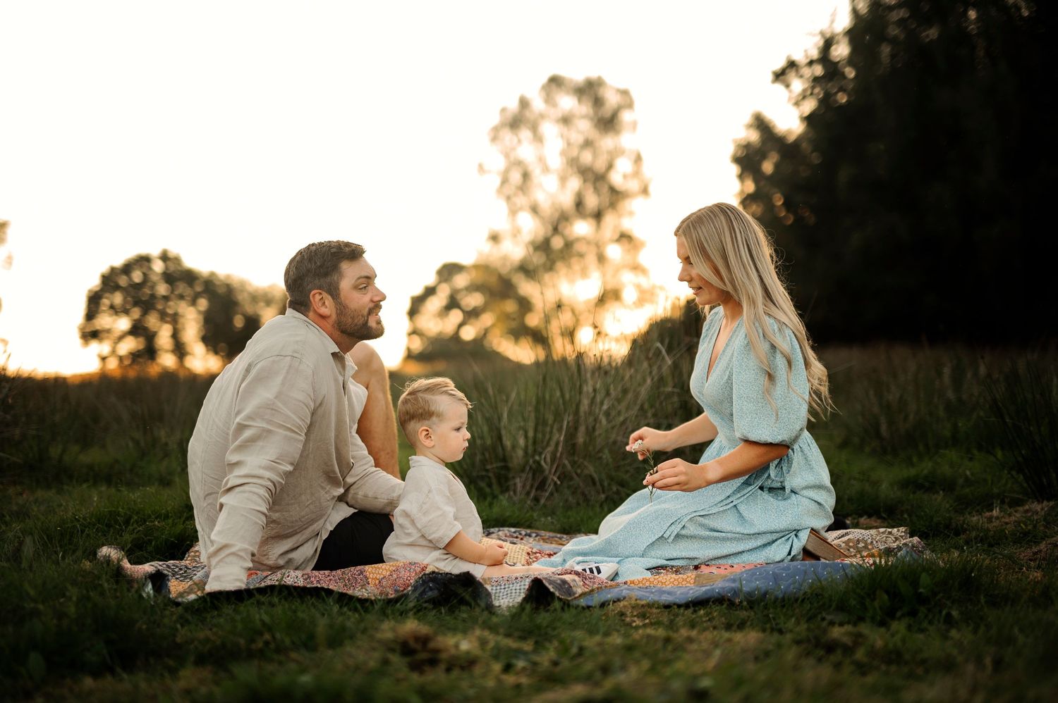 Family enjoying a picnic together on a blanket in a sunlit meadow at dusk.
