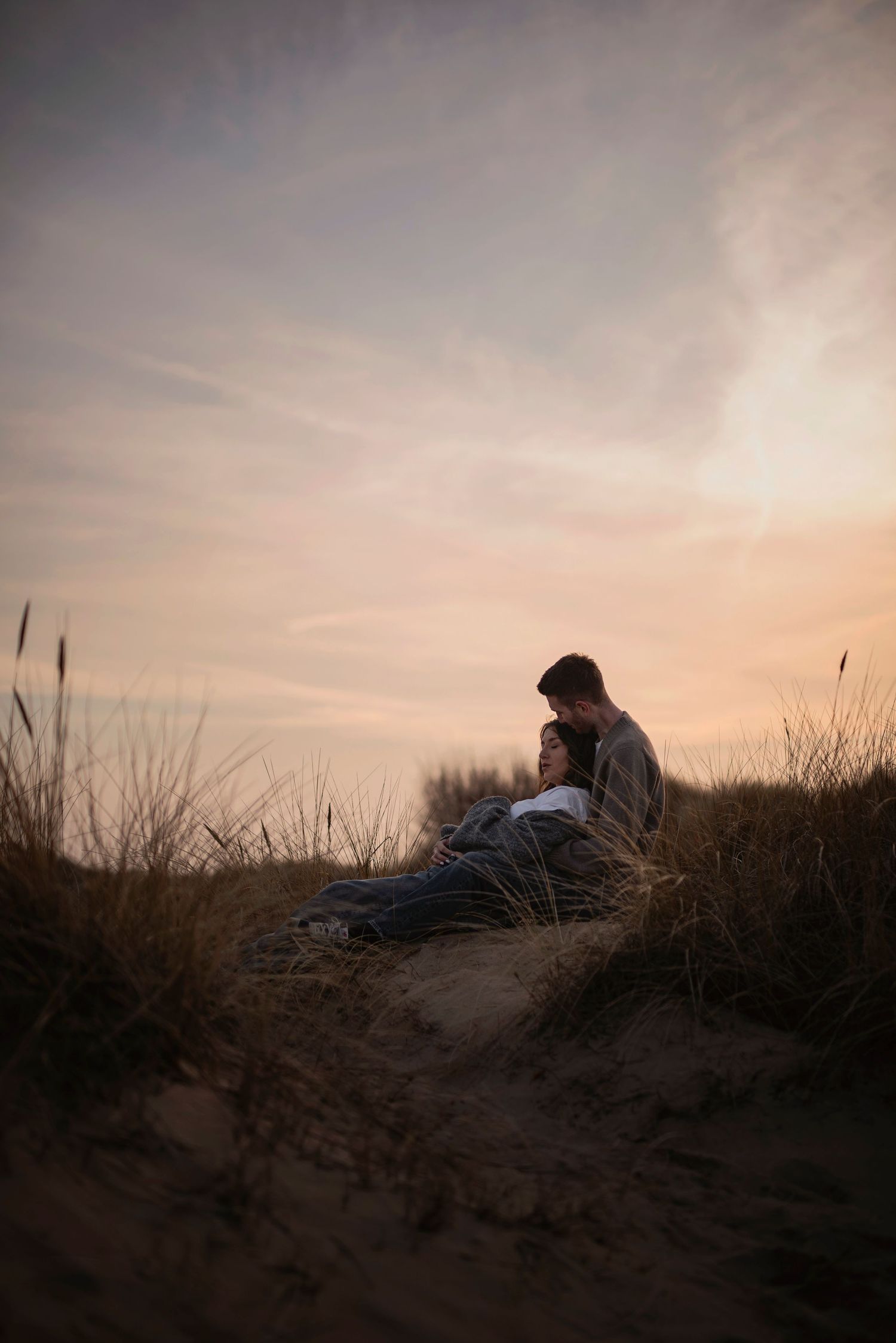 A silhouetted figure sits on a sandy dune at sunset while tall grasses sway in the gentle breeze.