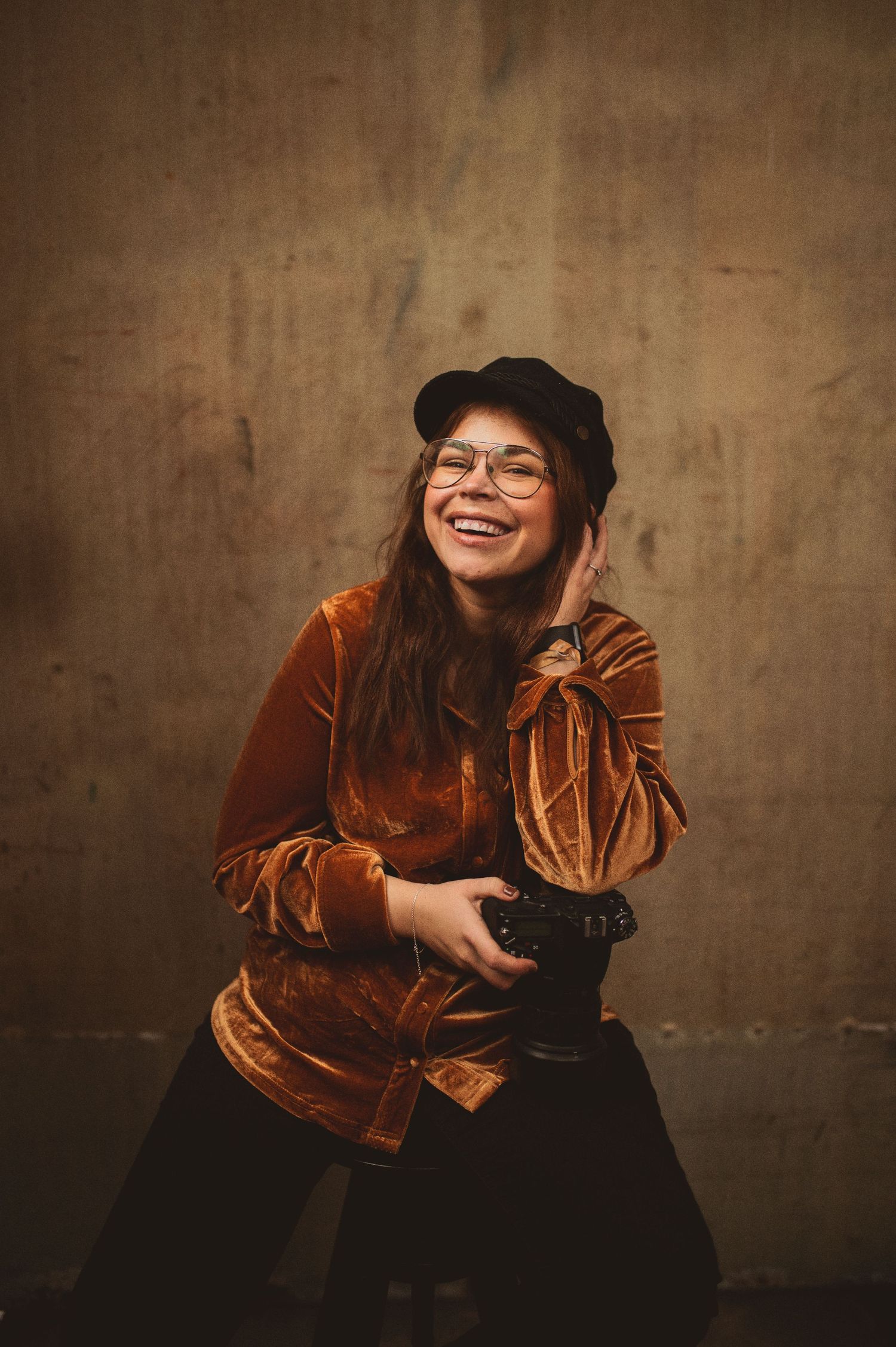 A smiling young woman wearing glasses and a black cap holds a camera while wearing a brown velvet jacket.