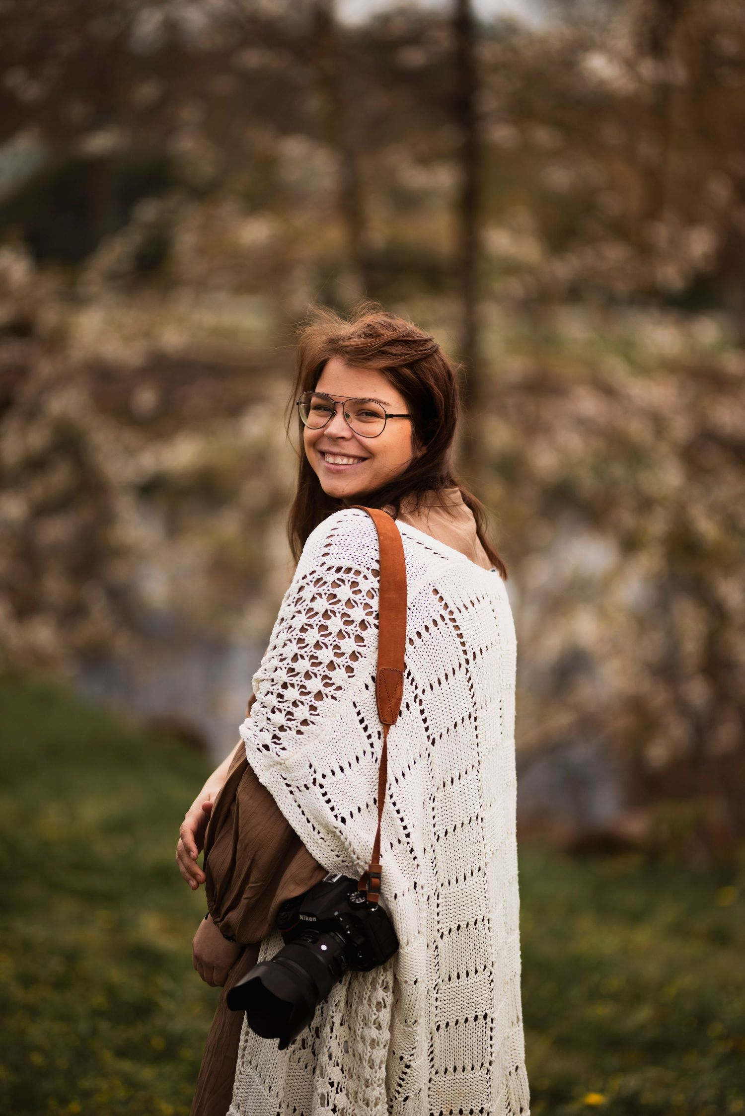 Smiling female photographer with glasses wearing white knit cardigan holding camera outdoors in autumn setting.