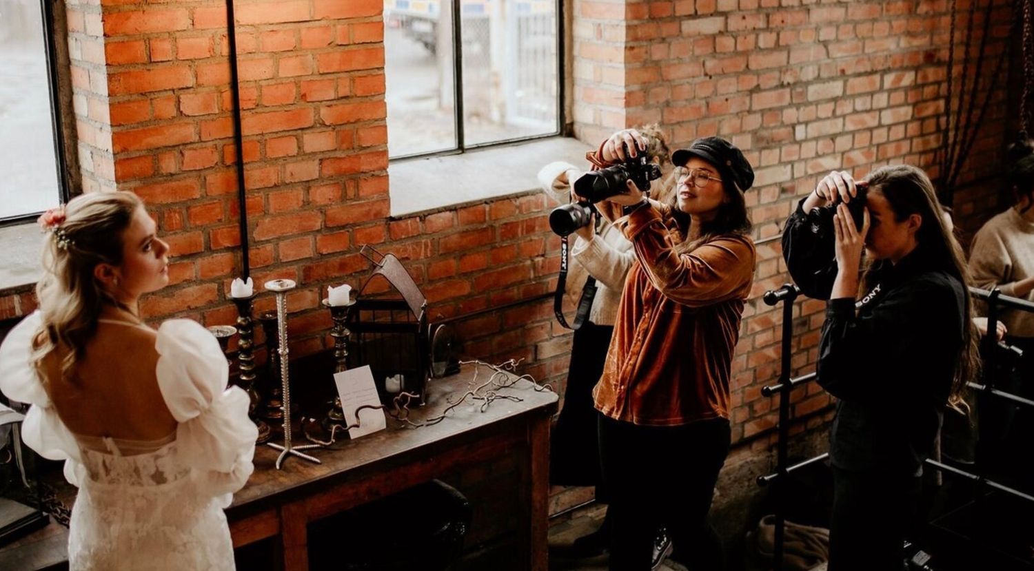 Multiple photographers shooting a model in a white dress inside a rustic brick-walled photography studio.