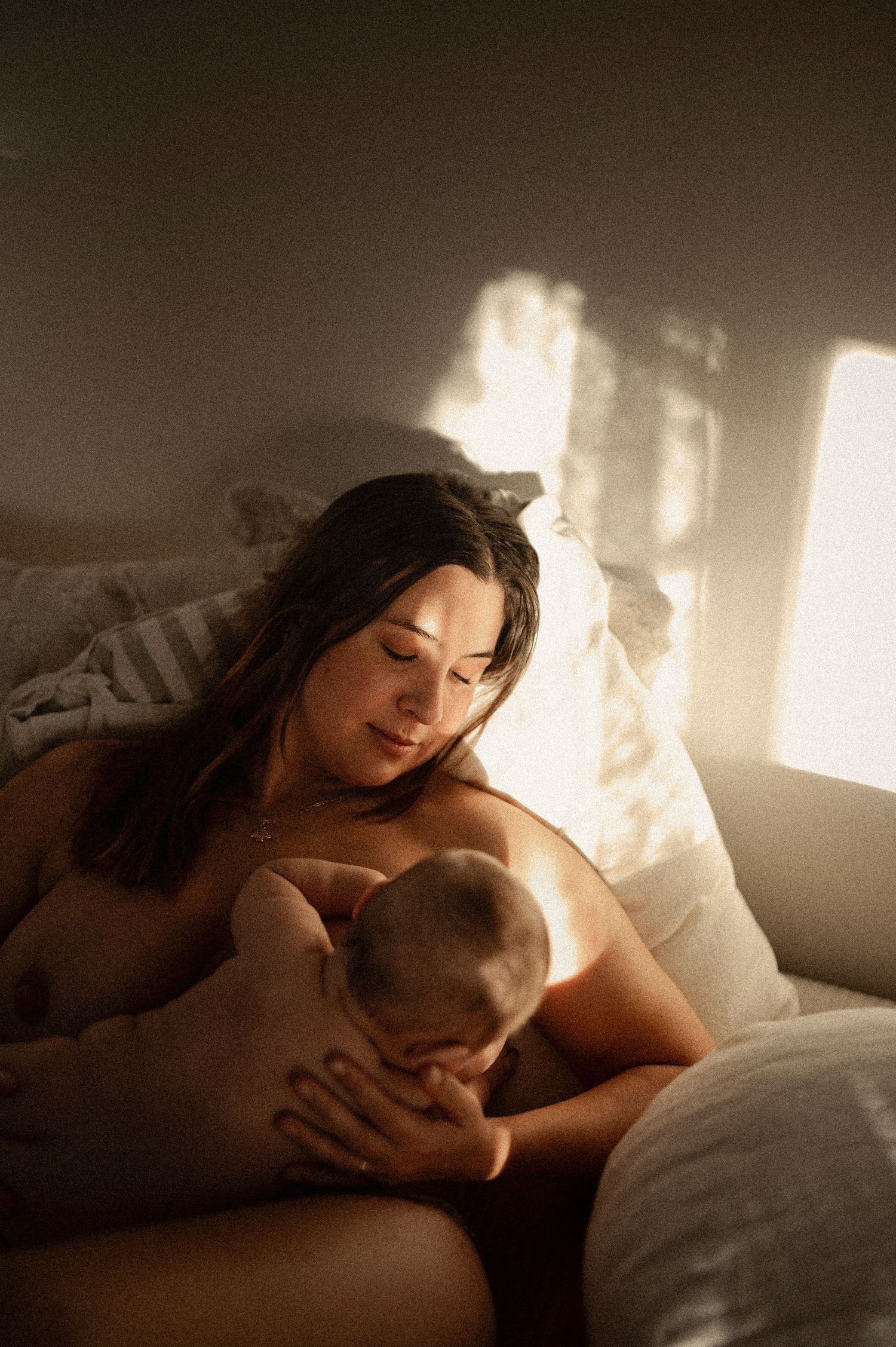 Mother breastfeeding her newborn baby in warm golden sunlight streaming through a window.