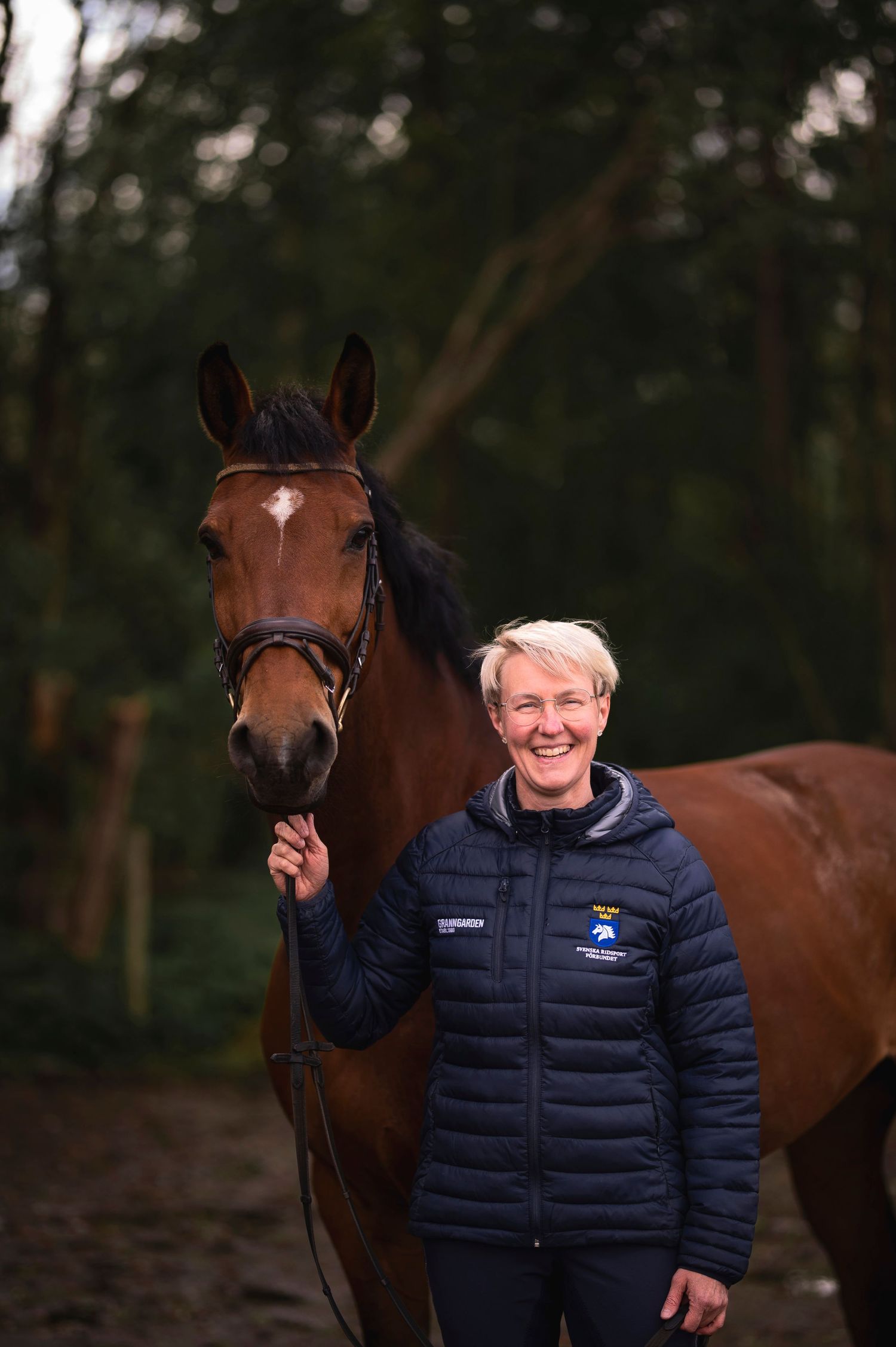 Happy woman in navy equestrian jacket standing beside a chestnut horse in a woodland environment.