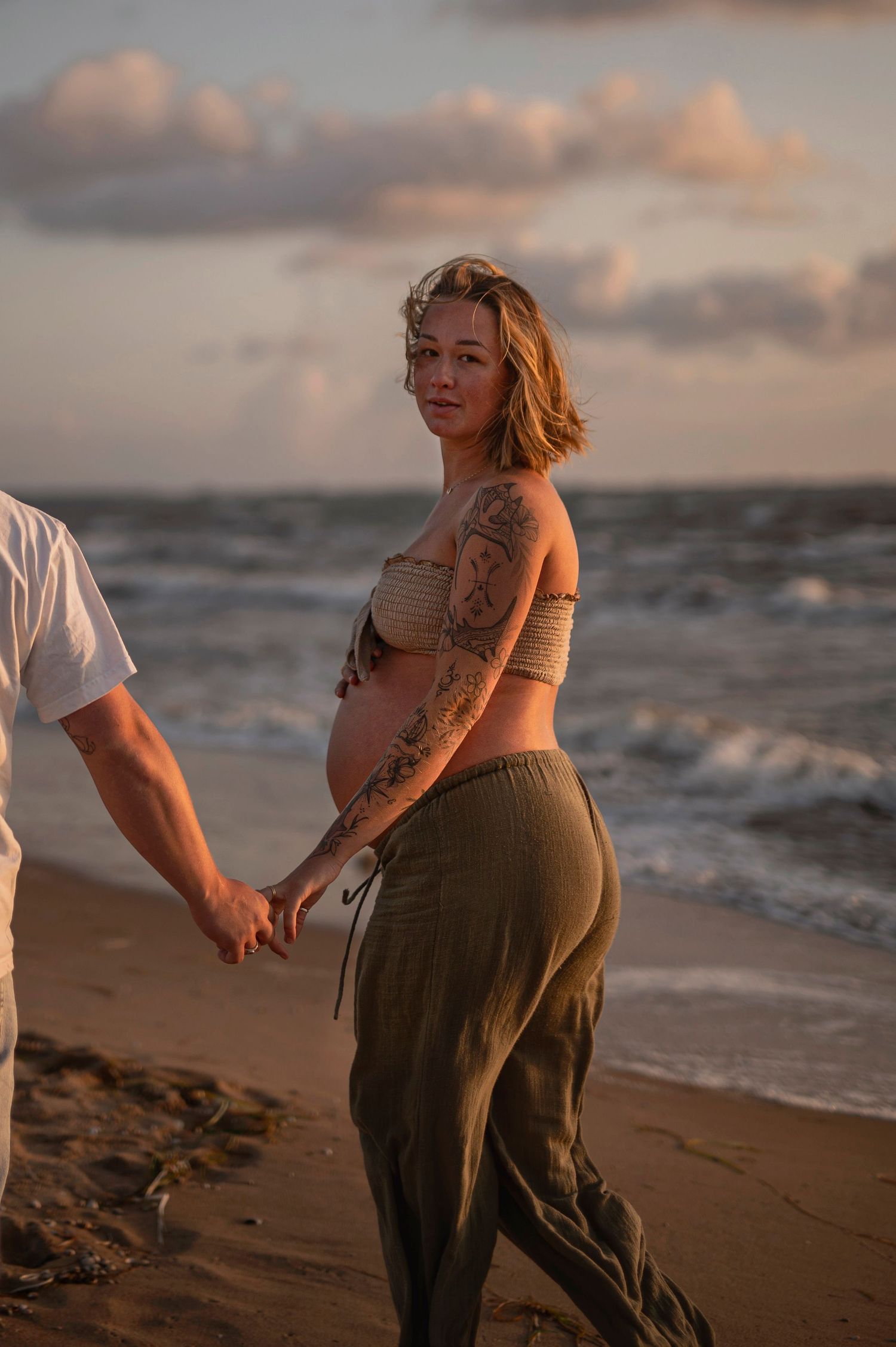 A pregnant woman in casual attire holds hands with someone during golden hour on a beach at sunset.