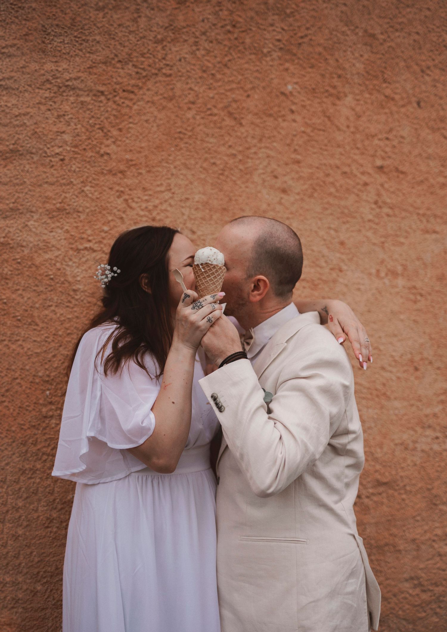 Newlyweds share a tender moment while eating ice cream against a terracotta wall.