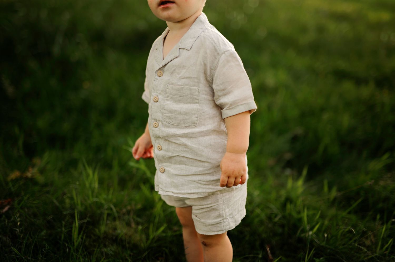 Small child wearing light colored outfit stands in lush green grass during sunset.