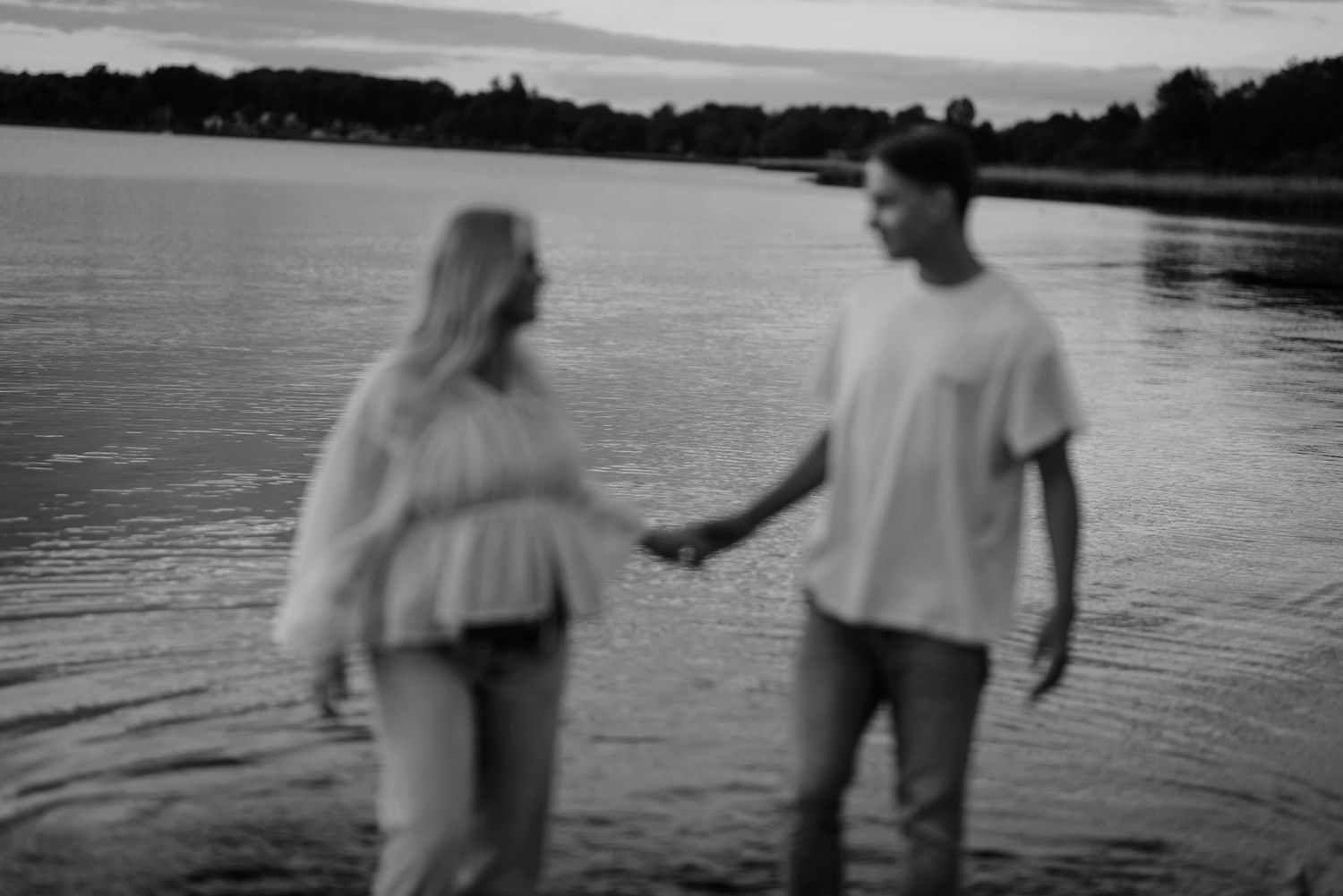 A blurry black and white image of a couple standing in shallow water at sunset by a lake.