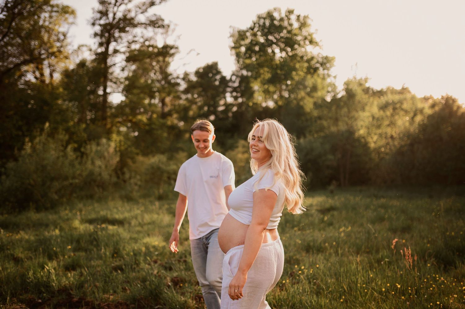 A romantic maternity photo shows a couple in white clothing enjoying a sunset moment in a grassy field.