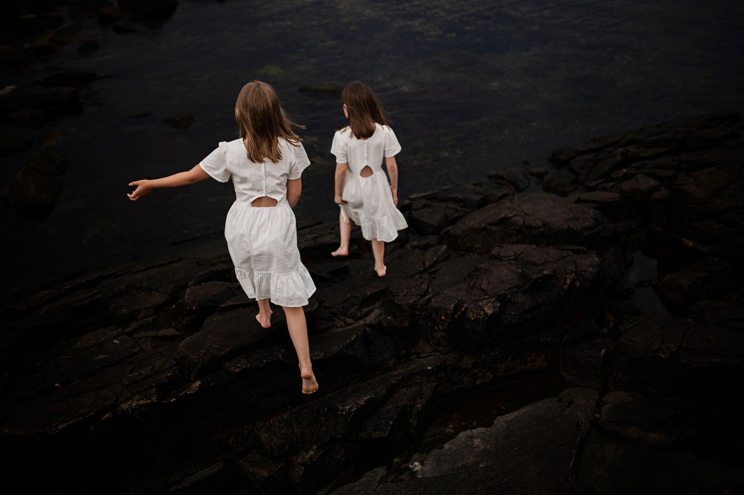 Two people in white dresses dance across dark rocky terrain.