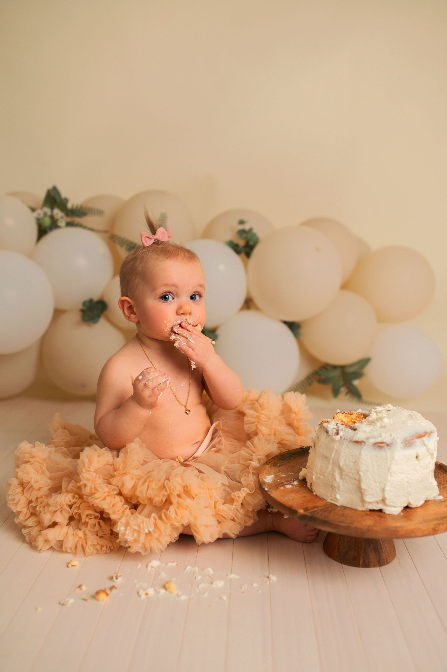 Baby in peach tulle skirt enjoying cake smash photo session with white balloons and greenery backdrop.