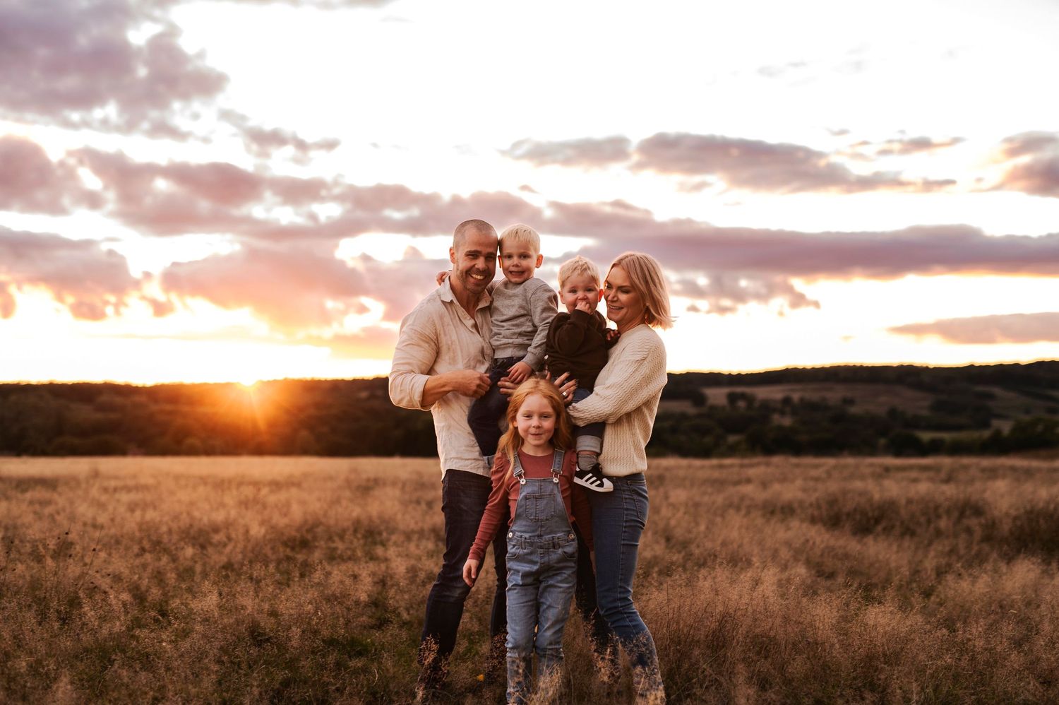 A loving family embraces while standing in a golden field at sunset.