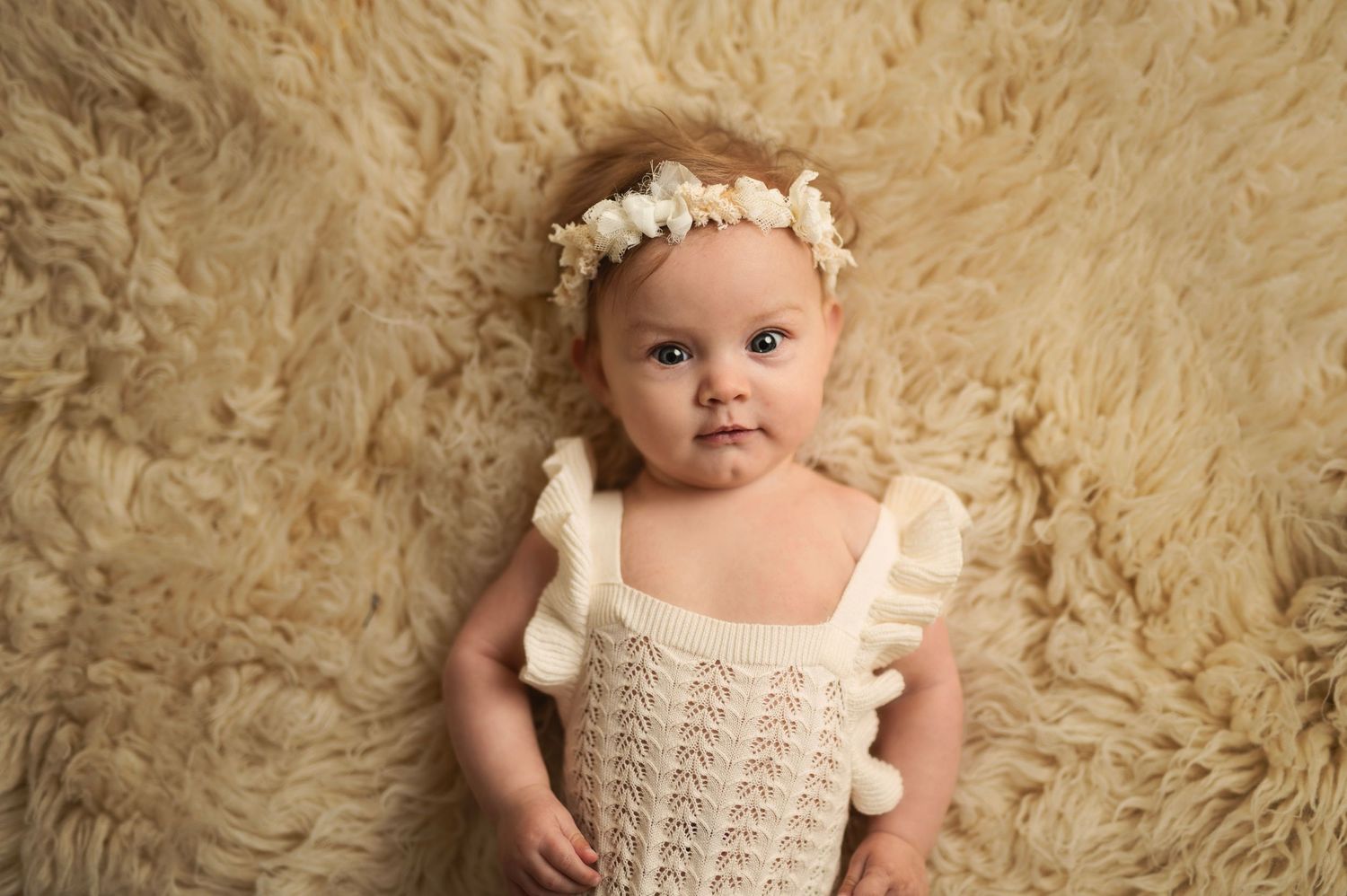 Baby wearing delicate white lace outfit and floral crown lying on plush beige fur background.