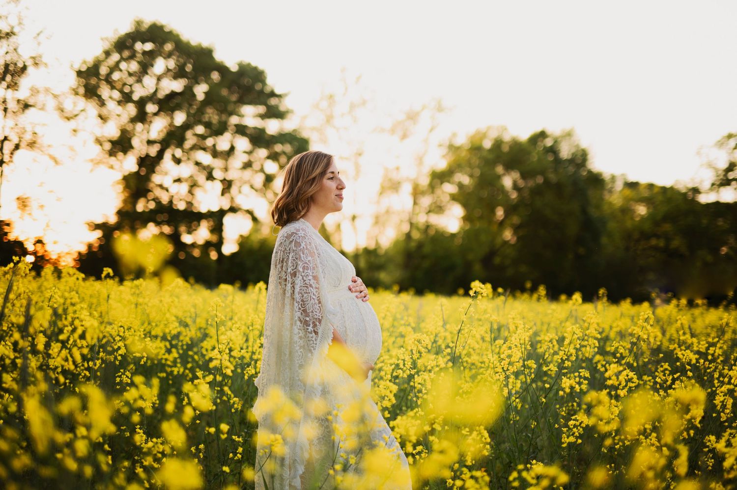 Ethereal maternity portrait of woman in white lace dress among yellow wildflowers.
