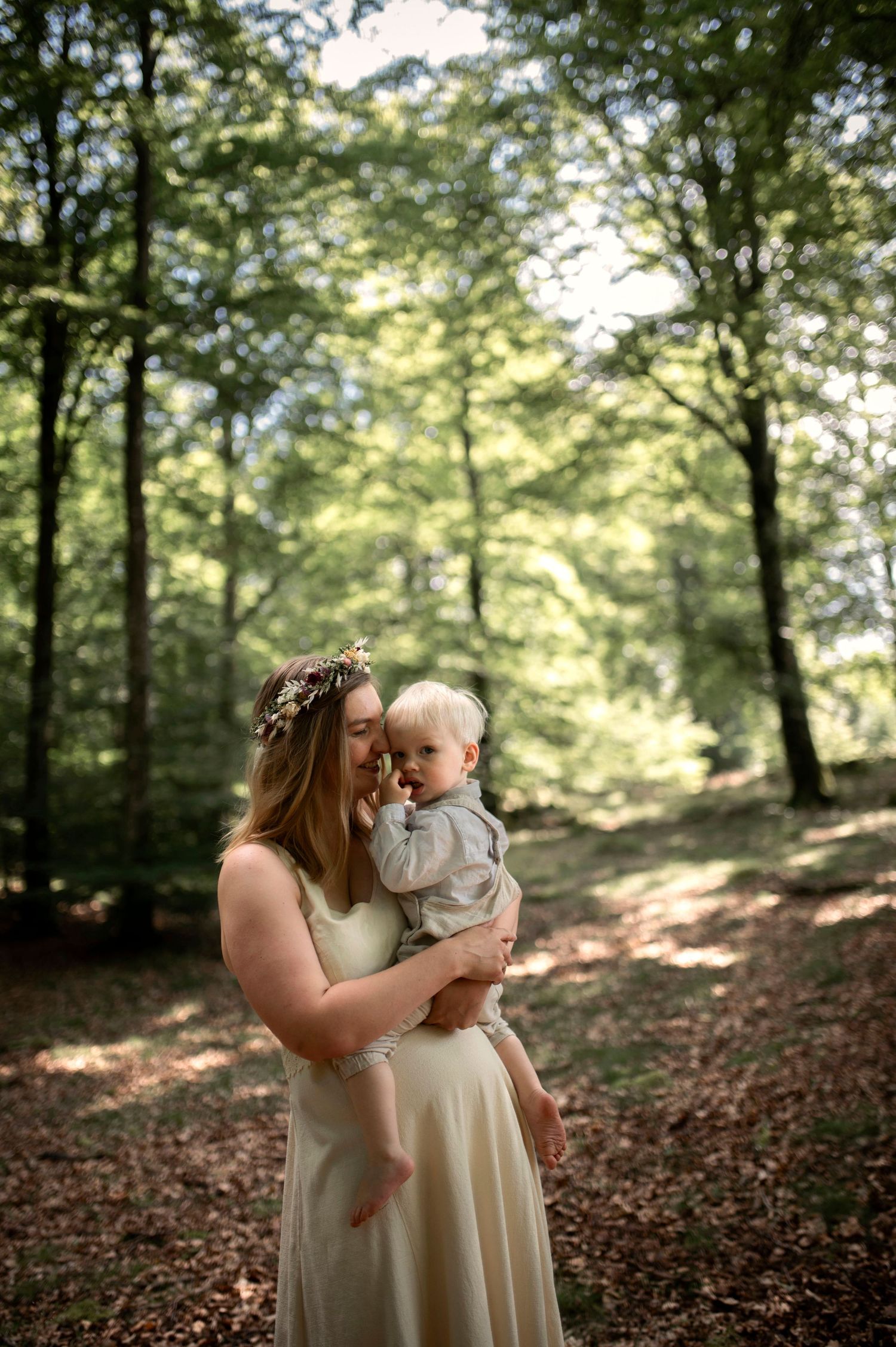 Mother wearing floral crown holding baby in sunlit forest clearing in cream dress.