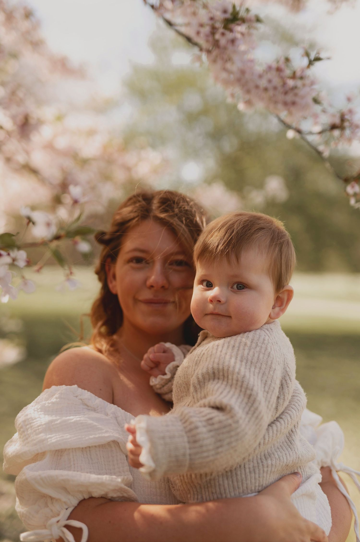 A mother holds her baby against a backdrop of soft pink cherry blossoms.