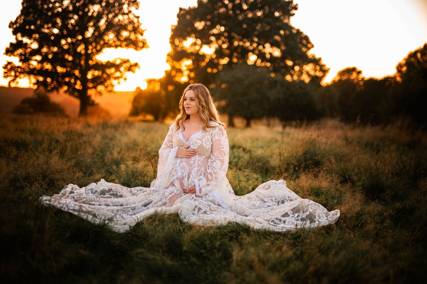 Serene maternity portrait with white lace gown spread across golden-lit field.