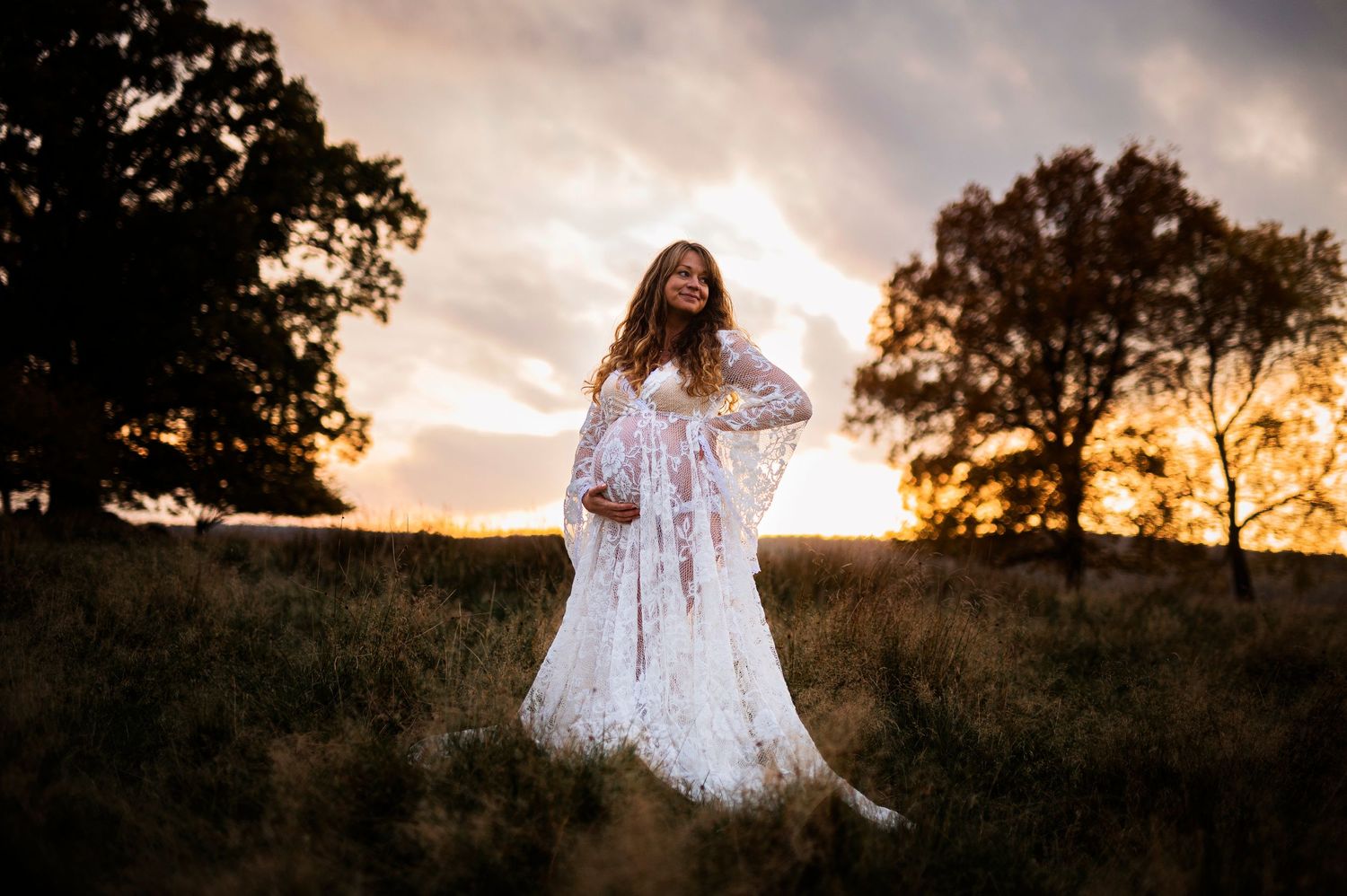 Dramatic maternity portrait in white dress against dusky sunset sky with trees.
