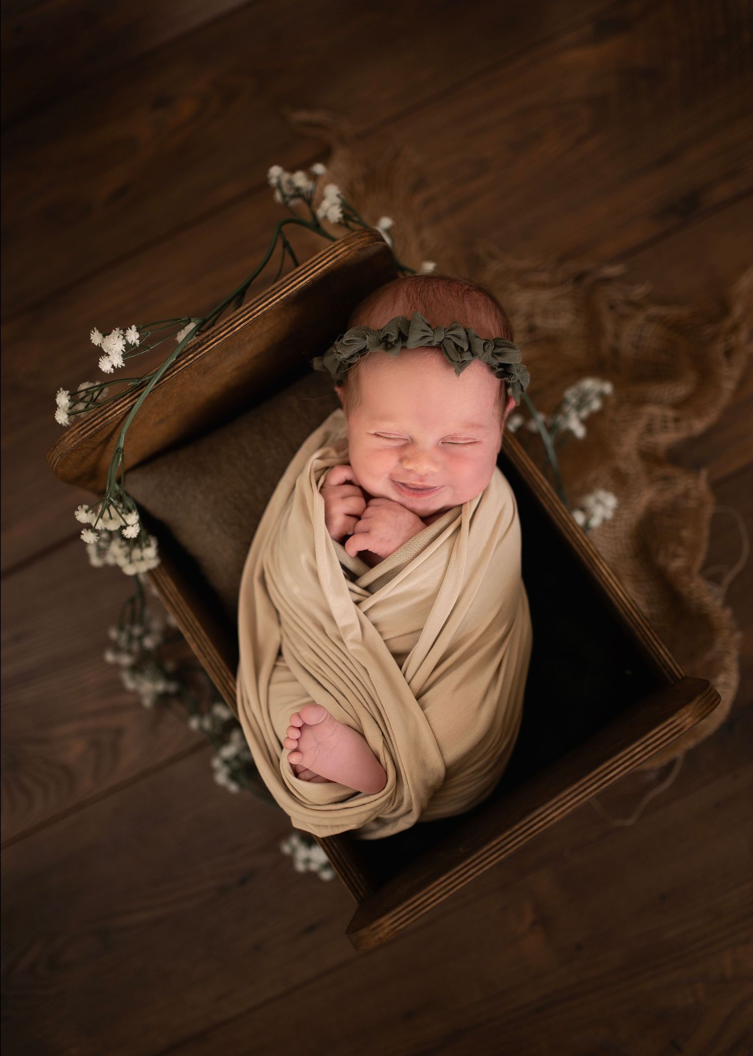Newborn photography with baby wrapped in beige fabric with floral crown in wooden box.