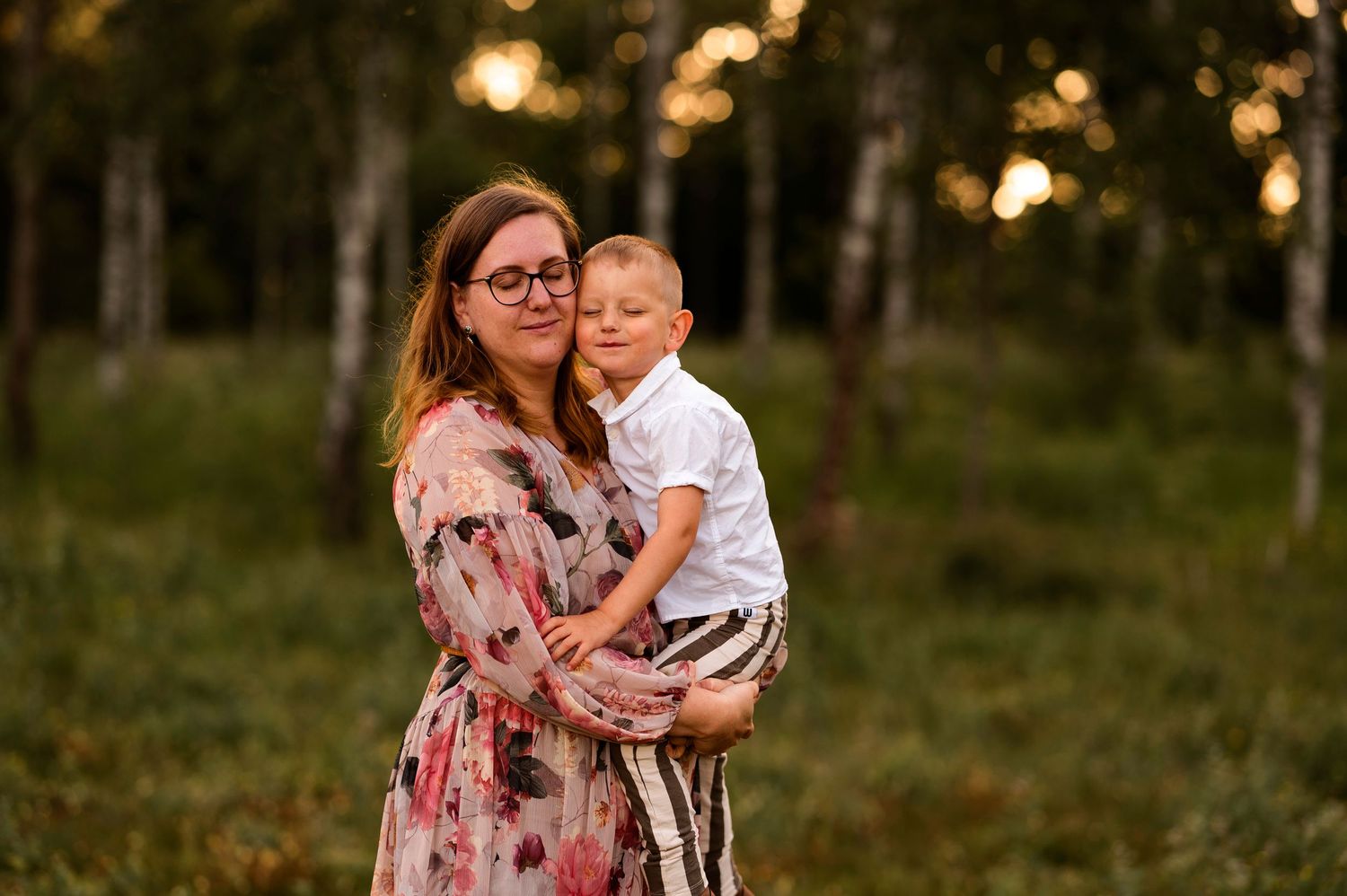 A warm family portrait in a floral dress against a forest backdrop at sunset.