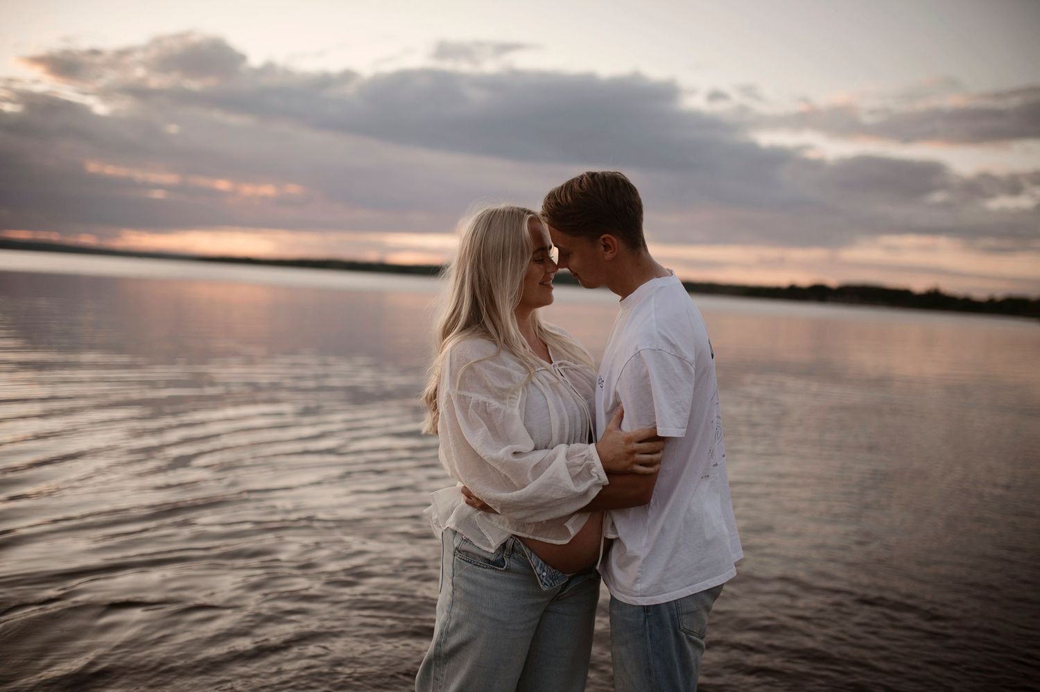 A tender moment between two people standing in shallow water at dusk with a peaceful lake backdrop.