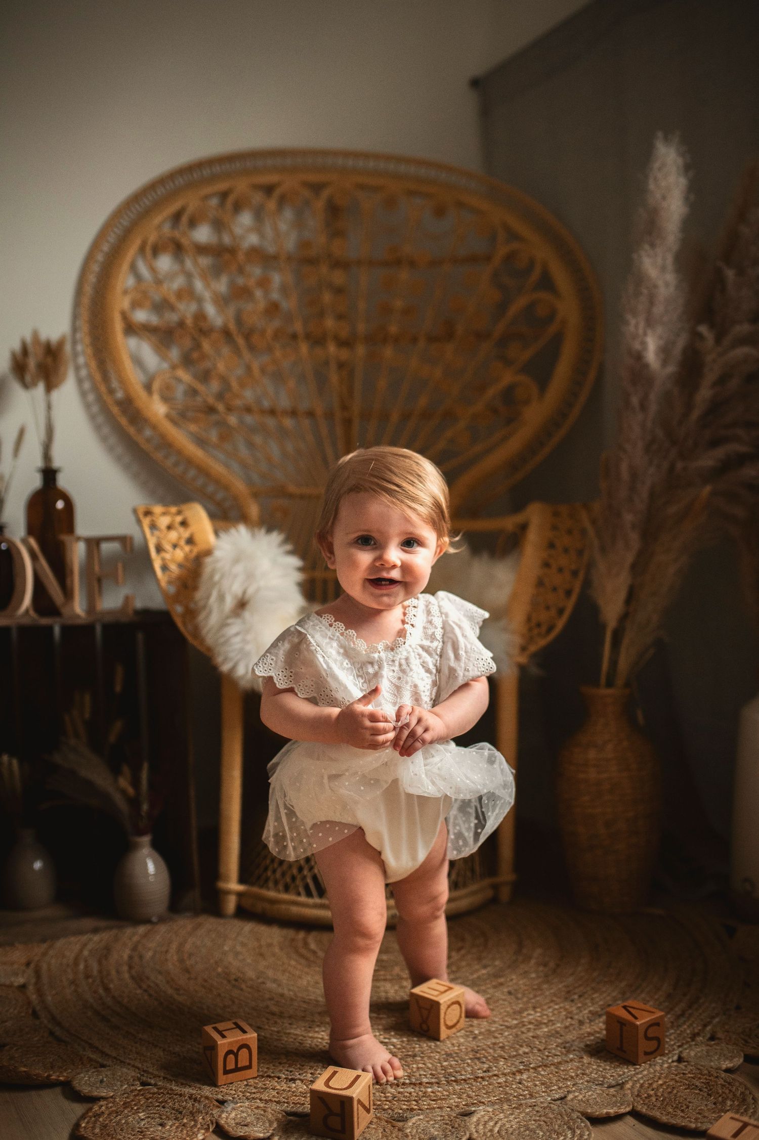 A small child in a white lacy dress plays with wooden blocks on a textured carpet near a vintage wicker peacock chair.