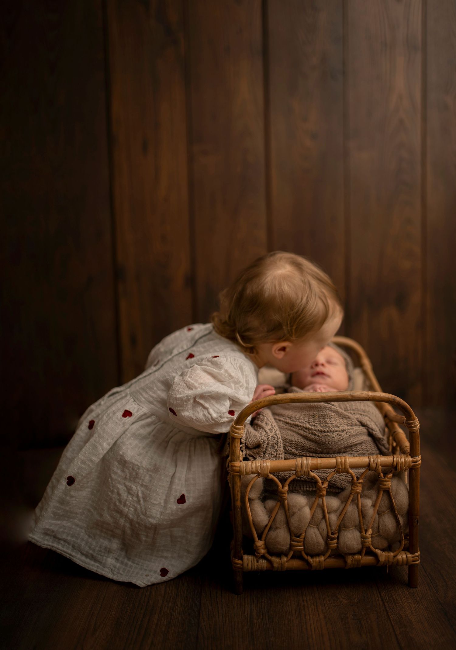 Sweet moment of toddler in white dress leaning over to kiss baby in wicker bassinet.