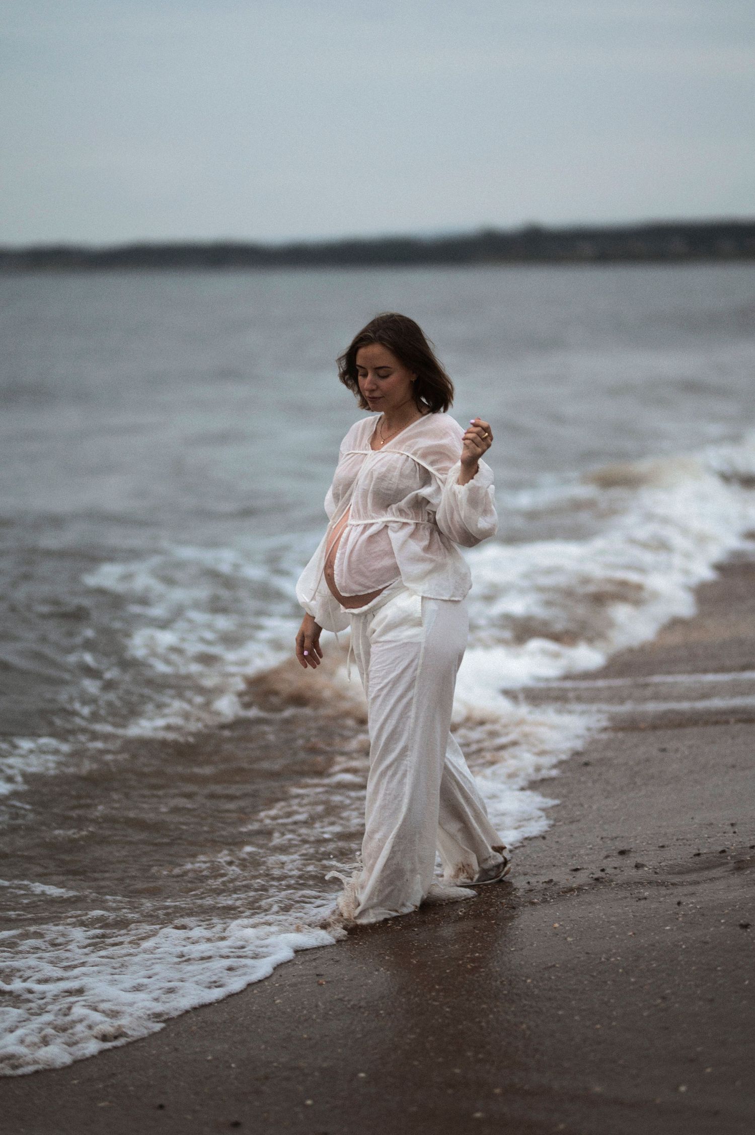 A pregnant woman in a flowing white dress stands at the edge of ocean waves on a moody beach.