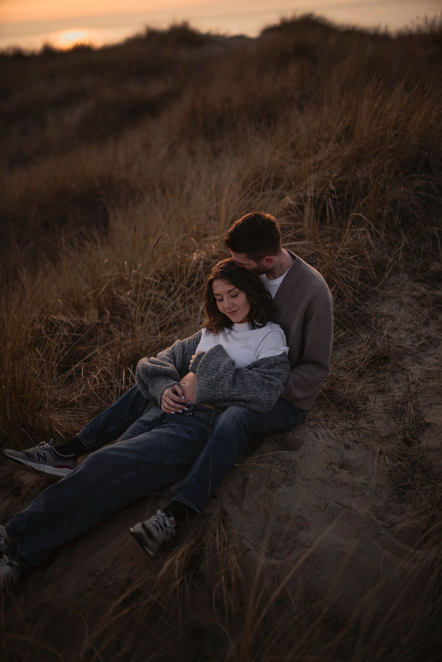 A tender moment between a couple resting on a sandy hill during golden hour at dusk.