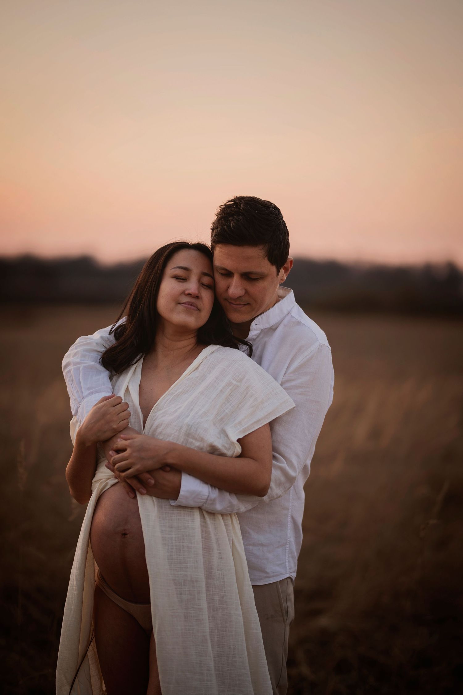 Intimate maternity photo of couple embracing in a field at sunset with soft warm lighting.