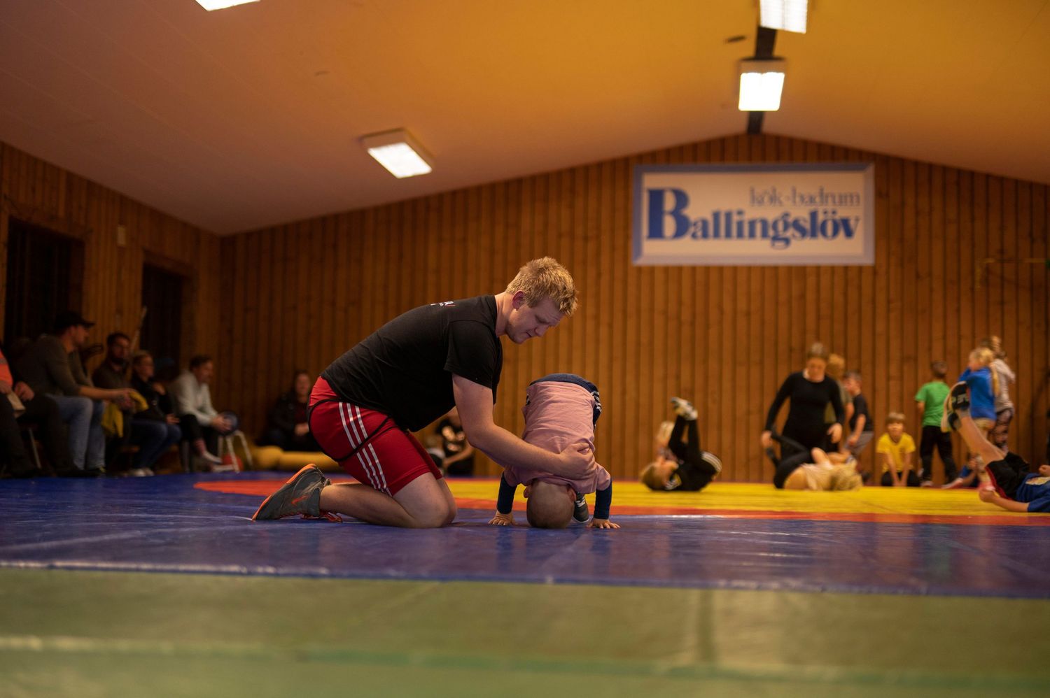 Young wrestler in red shorts practicing a takedown move on a blue mat in an indoor sports hall.