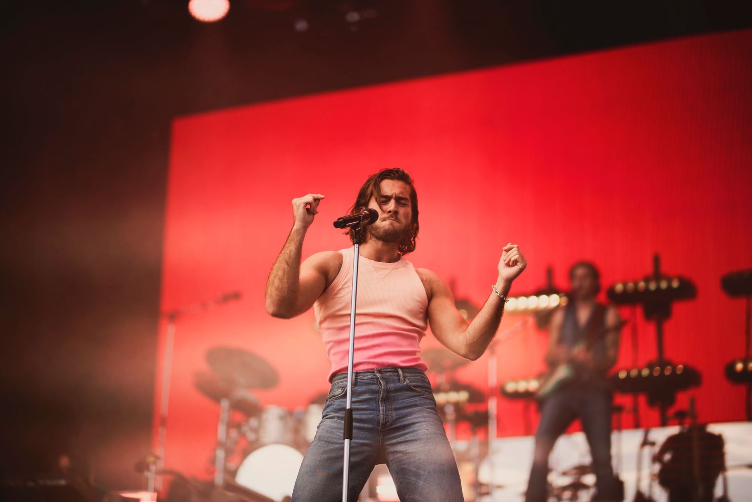 Energetic male performer in pink tank top singing on stage with red backdrop.