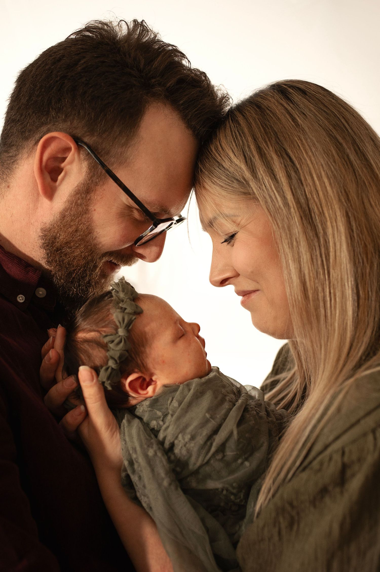 Loving parents gently holding sleeping newborn wrapped in sage green cloth together.