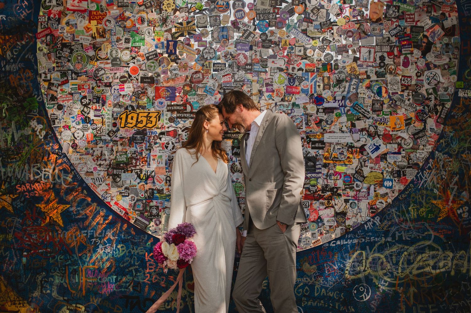 Couple embraces in front of blue heart graffiti wall holding purple wedding flowers.