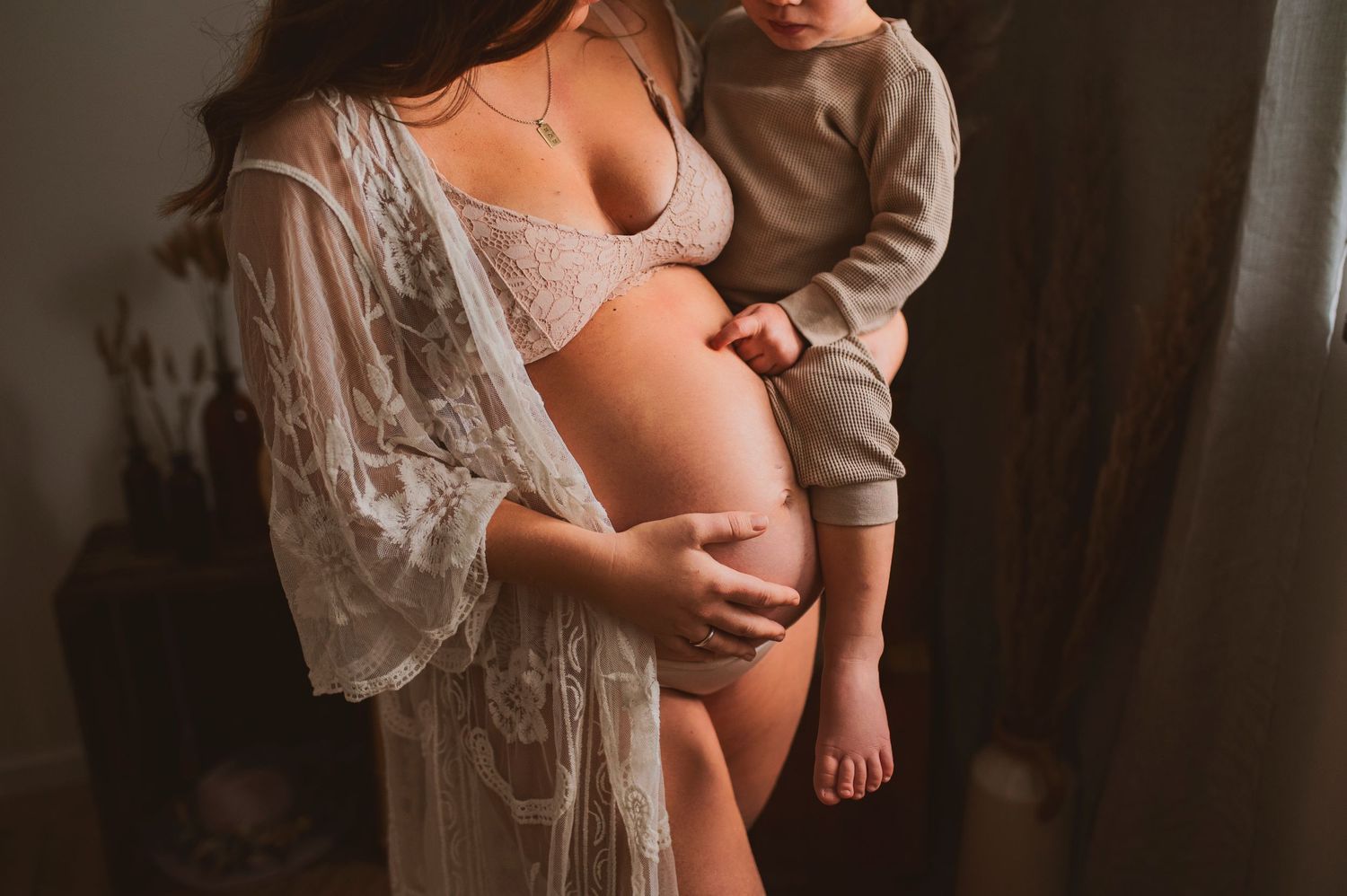 Intimate maternity photo showing a pregnant woman in white lace lingerie holding her belly with a child beside her.