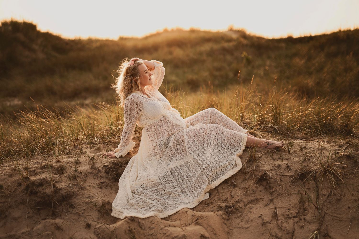 Ethereal maternity photo of figure in white lace dress reclining on sand dunes at golden hour.
