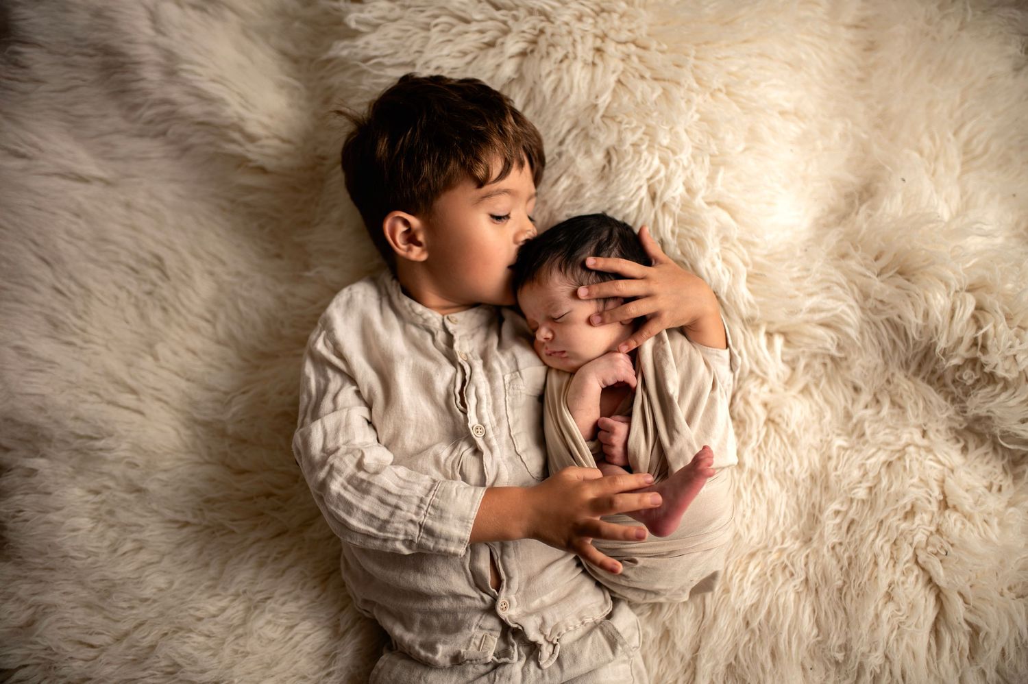 A sweet sibling moment captured on soft white fur as a young child gently kisses a newborn baby.